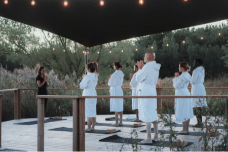 Groupe de personnes en peignoirs pratiquant le yoga sur une terrasse extérieure en bois, entourée de végétation naturelle, avec tapis de yoga au sol et éclairage suspendu créant une ambiance de détente.