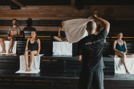 Rituel Aufguss dans un sauna en bois, avec un maître Aufguss utilisant une serviette pour diffuser la chaleur devant un groupe de personnes assises sur des bancs, profitant d’une expérience de bien-être thermique.