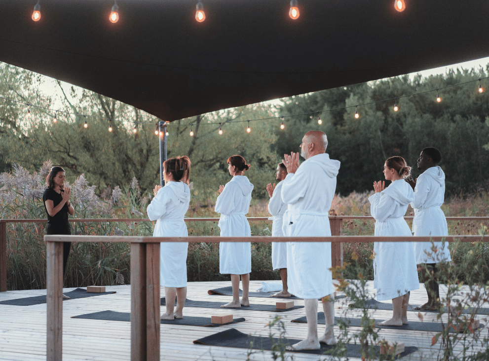 Group of people in bathrobes practicing yoga on an outdoor wooden terrace, surrounded by natural vegetation, with yoga mats on the floor and overhead lighting creating a relaxing atmosphere.