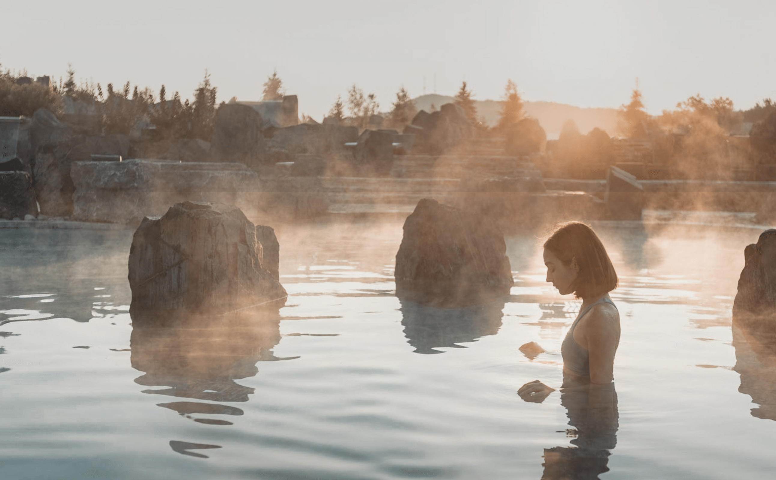 Personne immergée dans une source thermale extérieure entourée de rochers, au coucher du soleil, avec vapeur sur l’eau et paysage naturel en arrière-plan.