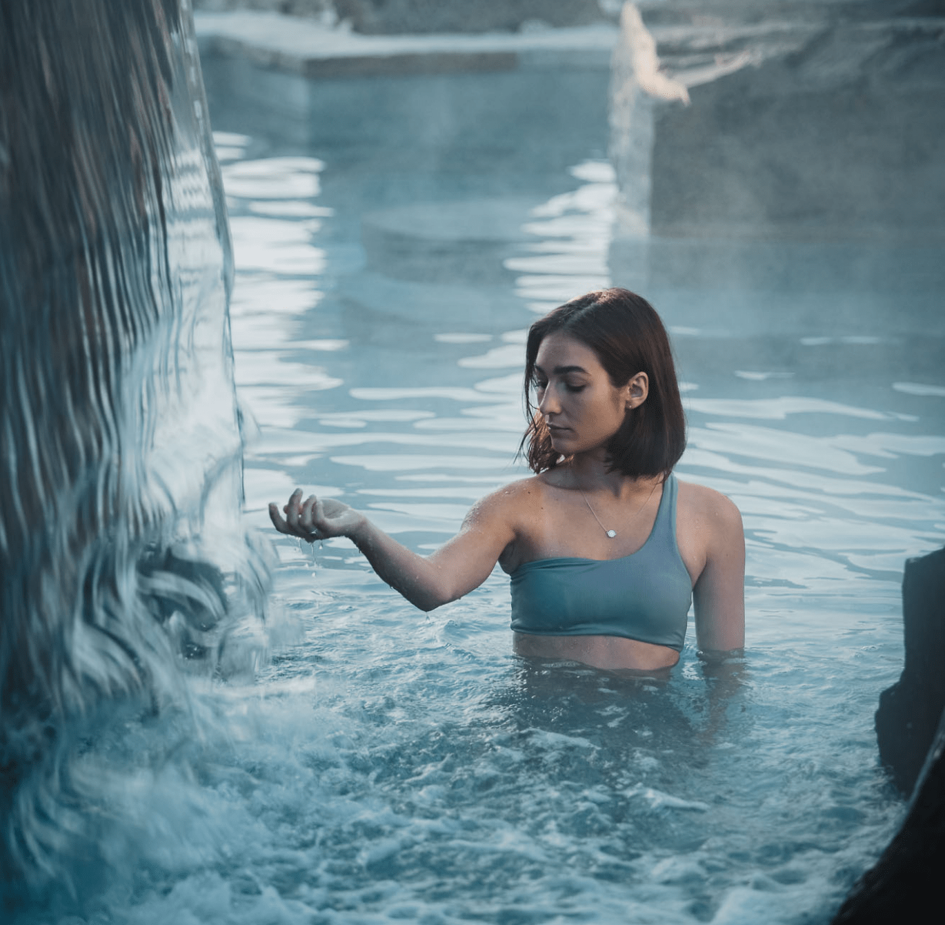 Personne dans un bassin thermal extérieur sous une chute d’eau, entourée de rochers, avec remous à la surface et ambiance spa naturelle apaisante.