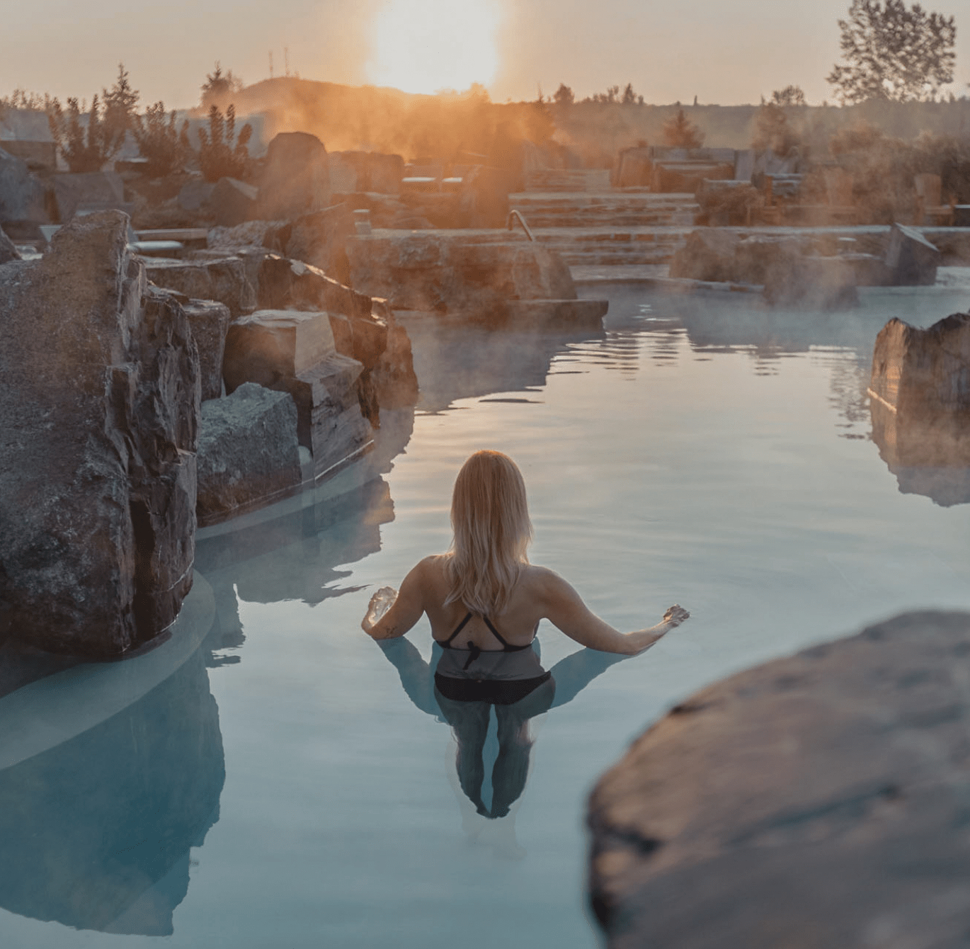 Personne de dos dans une source thermale extérieure entourée de rochers, au coucher du soleil, avec vapeur visible à la surface de l’eau dans un cadre naturel paisible.
