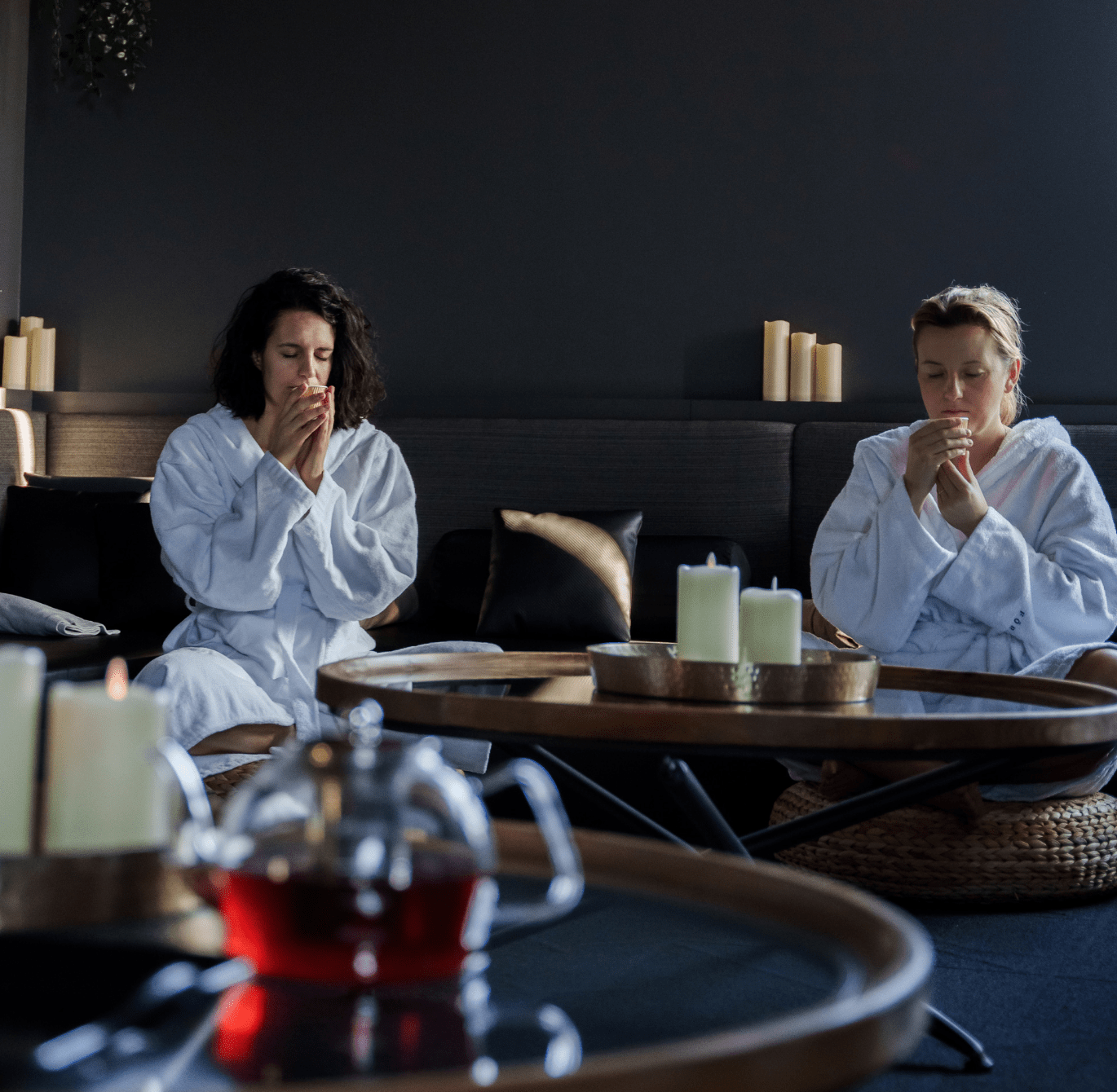 Deux personnes en peignoir blanc assises dans un espace spa intérieur tamisé, dégustant du thé lors d’un rituel du moment présent, avec théière en verre et bougies allumées créant une ambiance calme et méditative.