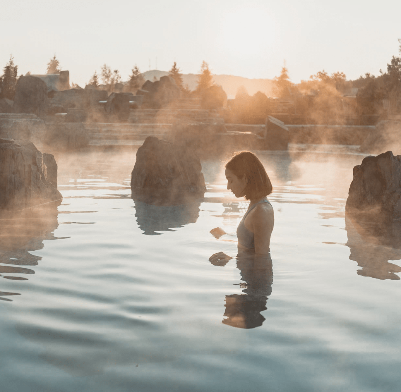 Personne immergée dans une source thermale extérieure entourée de rochers, au coucher du soleil, avec vapeur sur l’eau et paysage naturel en arrière-plan.