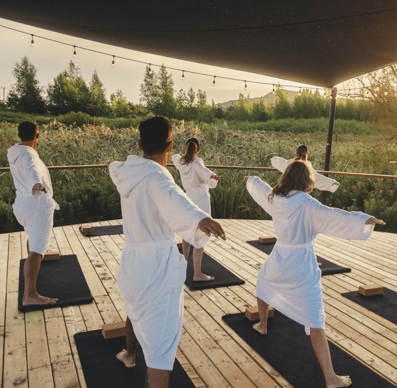 Groupe de personnes en peignoir blanc pratiquant une séance de yoga guidée sur une terrasse en bois extérieure, entourée de verdure, au coucher du soleil, avec tapis de yoga alignés et ambiance bien-être relaxante.